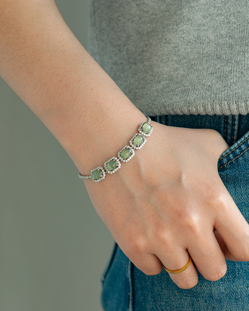 LYU_5678 A person wearing a grey top and blue jeans rests their hand in their pocket. A silver bracelet with five green gemstone settings and a thin gold ring are on their wrist and finger.