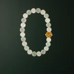 A beaded bracelet made of pale green and white round stones with a single ornate gold bead, displayed on a dark green background.