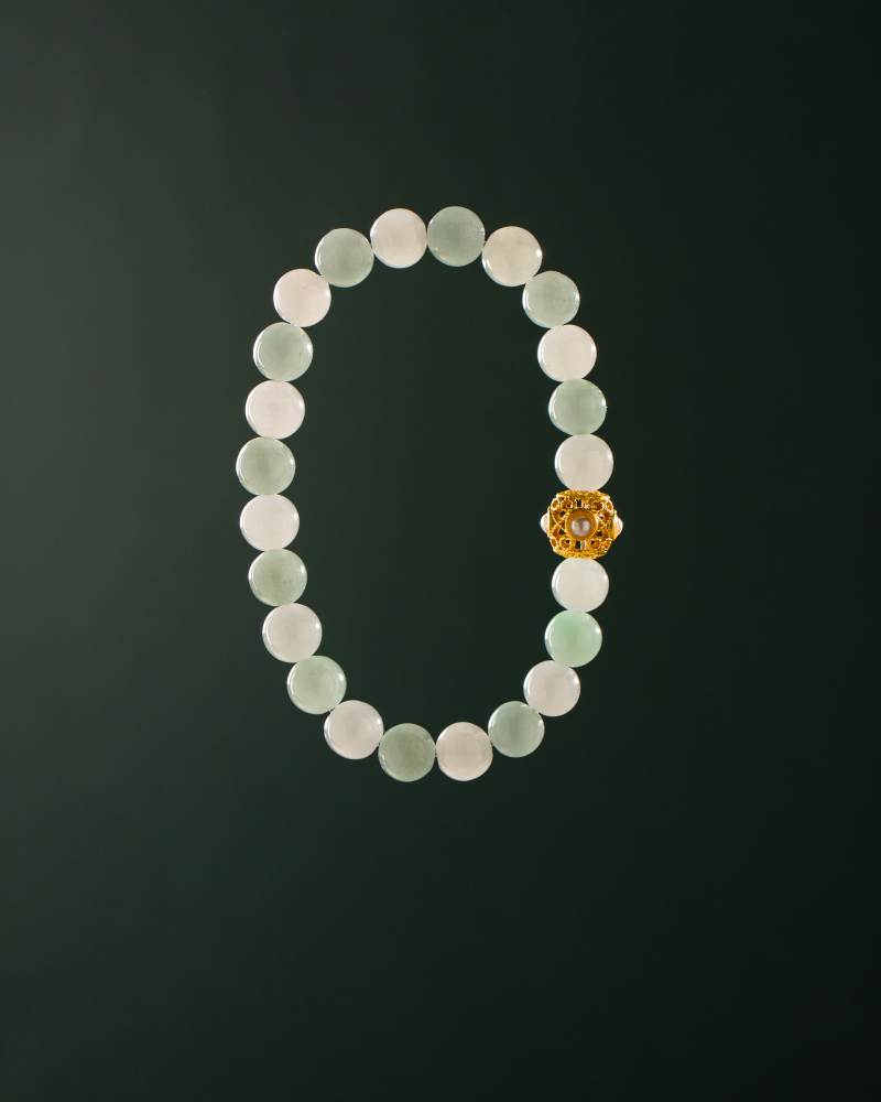 A beaded bracelet made of pale green and white round stones with a single ornate gold bead, displayed on a dark green background.