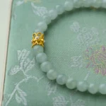 A jade bead bracelet with a gold floral clasp rests on a light green embroidered fabric box with a subtle floral pattern.