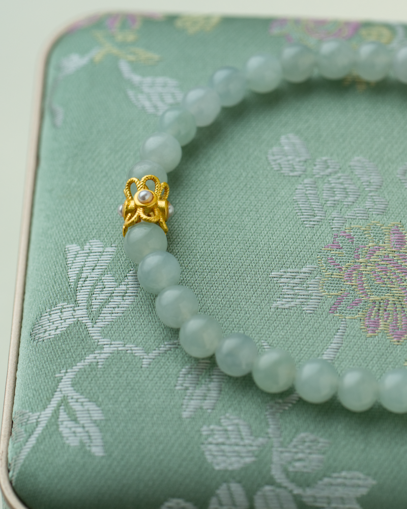 A jade bead bracelet with a gold floral clasp rests on a light green embroidered fabric box with a subtle floral pattern.