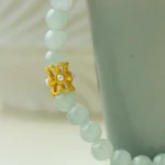 Close-up of a pale green beaded bracelet with a gold butterfly-shaped charm and a pearl detail, draped over the rim of a light green ceramic cup.