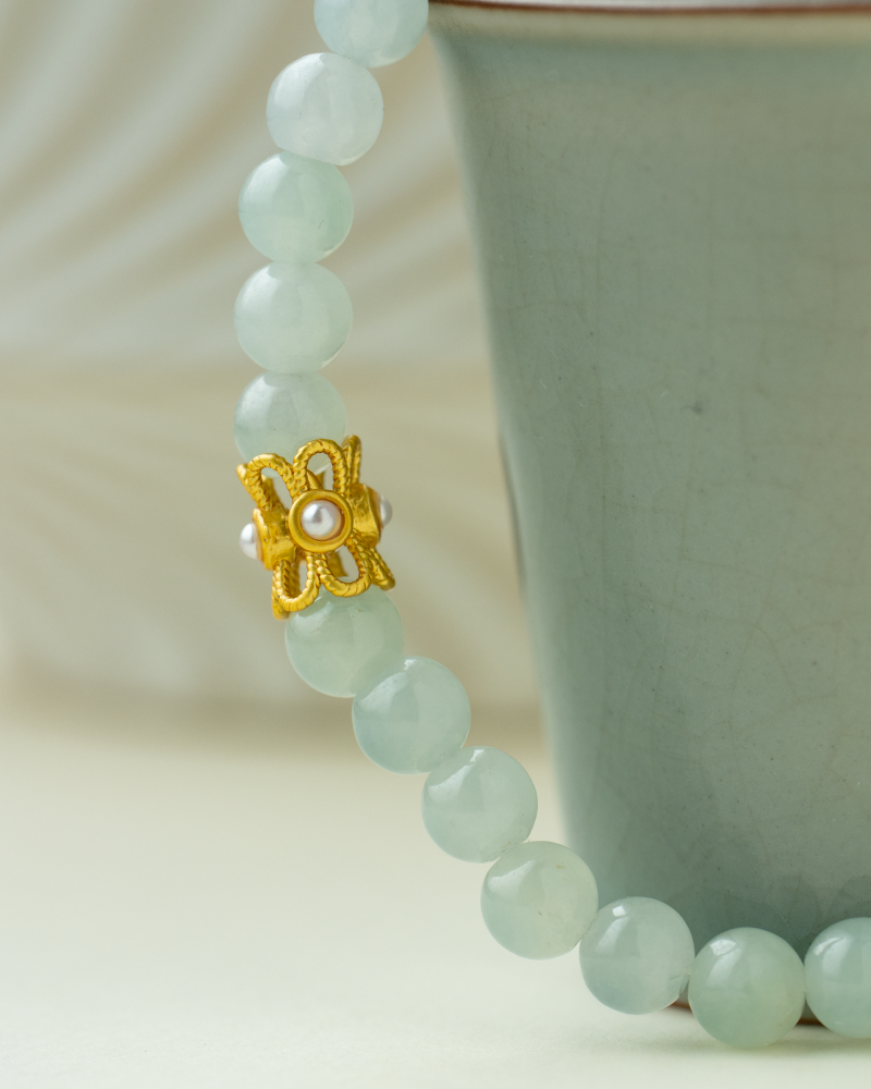 Close-up of a pale green beaded bracelet with a gold butterfly-shaped charm and a pearl detail, draped over the rim of a light green ceramic cup.