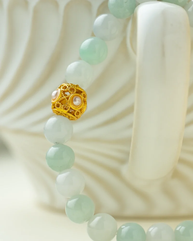 A close-up of the Light Green and Greenish-White Jadeite Bracelet—crafted with spherical beads and finished with a gold-tone accent detail—draped over the handle of a white, textured ceramic cup.