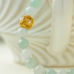 A close-up of a beaded necklace with pale green round beads and a single ornate gold bead, draped over the curved handle of a white, wavy-patterned ceramic cup.