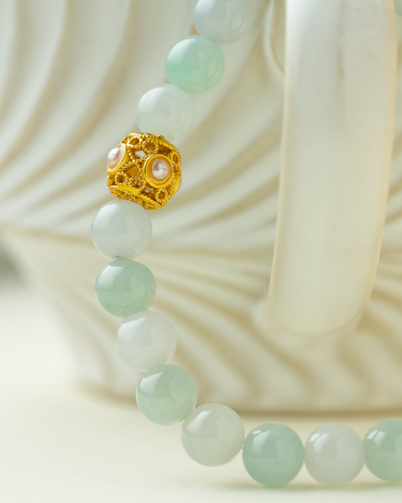 A close-up of a beaded necklace with pale green round beads and a single ornate gold bead, draped over the curved handle of a white, wavy-patterned ceramic cup.