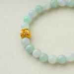 A beaded bracelet made of pale green jade stones with a single ornate gold and pearl bead, displayed on a light beige fabric background.