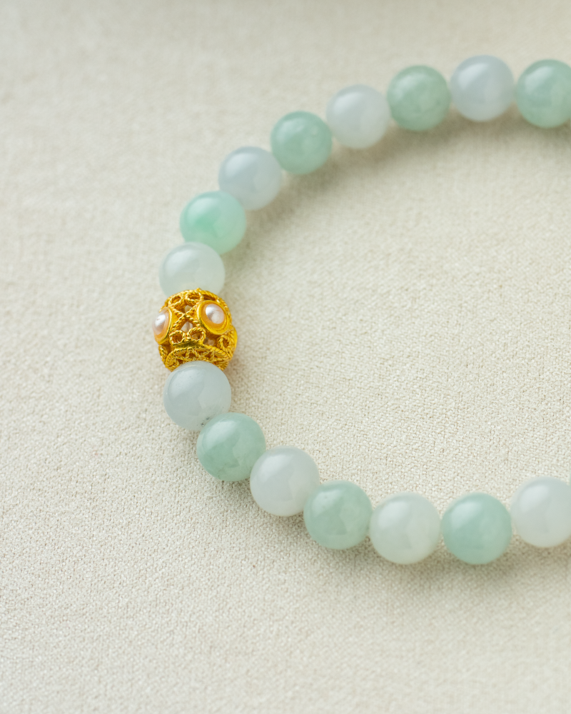 A beaded bracelet made of pale green jade stones with a single ornate gold and pearl bead, displayed on a light beige fabric background.