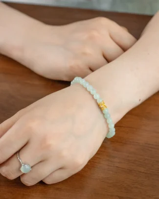 A Light Green Jadeite Bracelet — Spherical Beads with Gold-Tone Accent Detail is worn on the wrist. The hand also features a delicate silver ring set with a light green stone. Both hands are resting on a wooden surface.