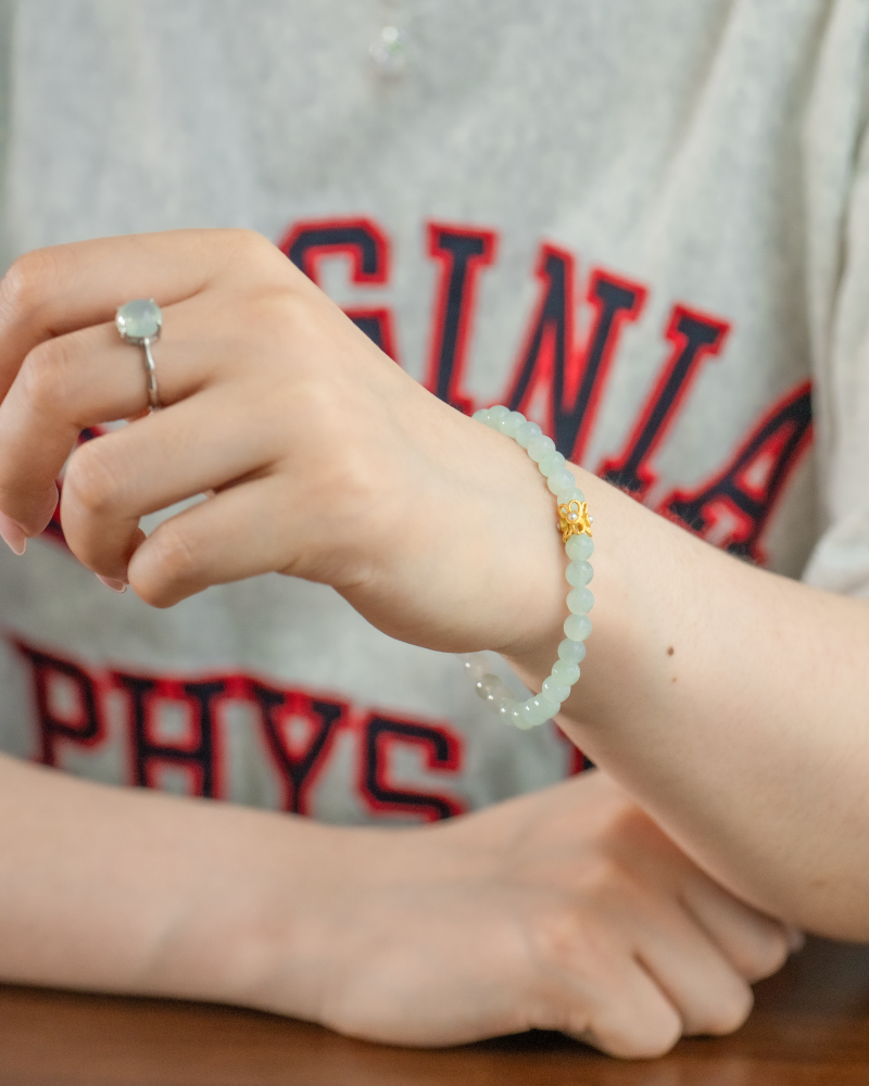 A person wearing a gray T-shirt with red letters, a beaded bracelet with a gold charm, and a silver ring with a light blue stone on their finger, resting their hand on a table.