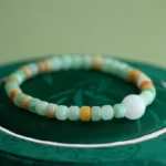 A beaded bracelet with green, white, and amber-colored stones rests on a dark green, textured surface with a soft, blurred green background.