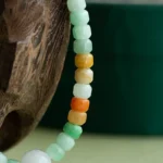 Close-up of a beaded necklace with square and round jade beads in green, yellow, amber, and orange hues, draped over a textured wooden surface with a blurred green background.
