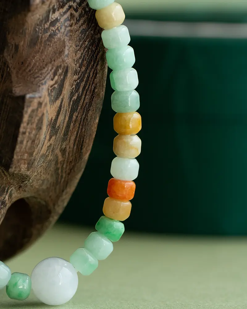 DSC_0549 拷贝 Close-up of a beaded necklace with square and round jade beads in green, yellow, amber, and orange hues, draped over a textured wooden surface with a blurred green background.