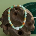A beaded bracelet with light green, yellow, orange, and white round beads is displayed on a carved wooden stand against a green background.