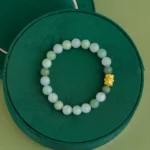 A green beaded bracelet with a single gold decorative bead is displayed on a round, dark green textured box, set against a light green background.