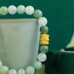 A jade beaded bracelet with a gold decorative bead is displayed draped over a light green ceramic cup, with a dark green circular box in the background.