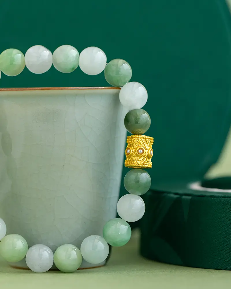 DSC_0552 拷贝 A jade beaded bracelet with a gold decorative bead is displayed draped over a light green ceramic cup, with a dark green circular box in the background.