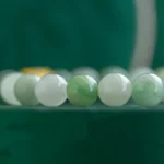 Close-up of a beaded bracelet featuring round jade beads in varying shades of green and white, set against a dark green background.