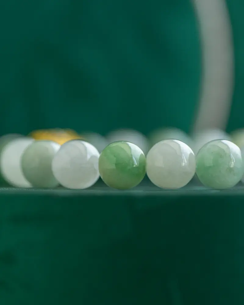 DSC_0553 拷贝 Close-up of a beaded bracelet featuring round jade beads in varying shades of green and white, set against a dark green background.