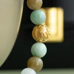 A close-up of a beaded bracelet featuring jade-green, white, brown, and a single engraved gold bead, draped over the edge of a smooth, light-colored surface with a blurred black and gold background.