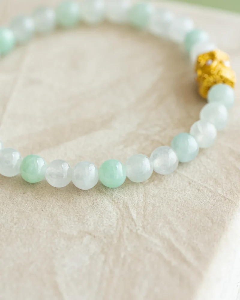 A close-up of the Light Green and White Jadeite Bead Bracelet—featuring spherical light green and white translucent beads with a single gold-tone openwork accent—resting on a beige textured surface.