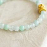 A close-up of a beaded bracelet featuring pale green and white jade beads with a single ornate gold bead, placed on a textured beige surface.