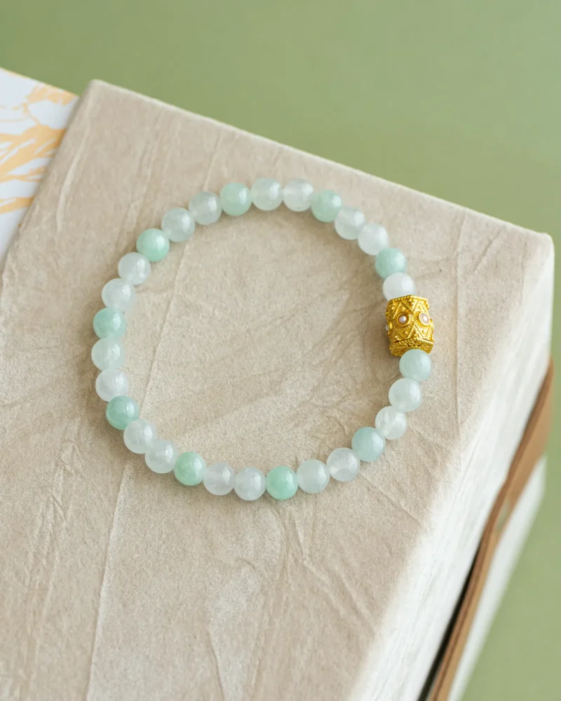 The Light Green and White Jadeite Bead Bracelet, crafted with spherical beads and highlighted by a gold-tone openwork accent, rests elegantly atop a textured beige box.