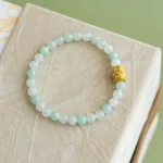 A beaded bracelet made of light green and white round stones with a single ornate gold bead, displayed on a beige textured surface.
