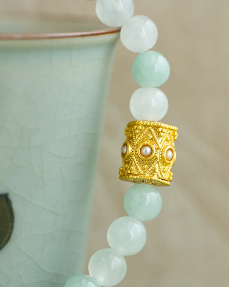 A close-up of the Light Green and White Jadeite Bead Bracelet—featuring spherical pale green and white jadeite beads and a gold-tone openwork accent bead—draped elegantly over the rim of a pale green ceramic cup.