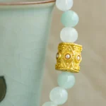Close-up of a beaded bracelet with pale green round beads and a detailed gold cylindrical bead with pearl-like accents draped over the rim of a light green ceramic cup.