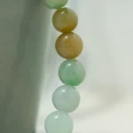 A close-up of a beaded jade bracelet draped over the edge of a light green ceramic cup, with beads in soft green and yellowish-brown hues on a neutral background.