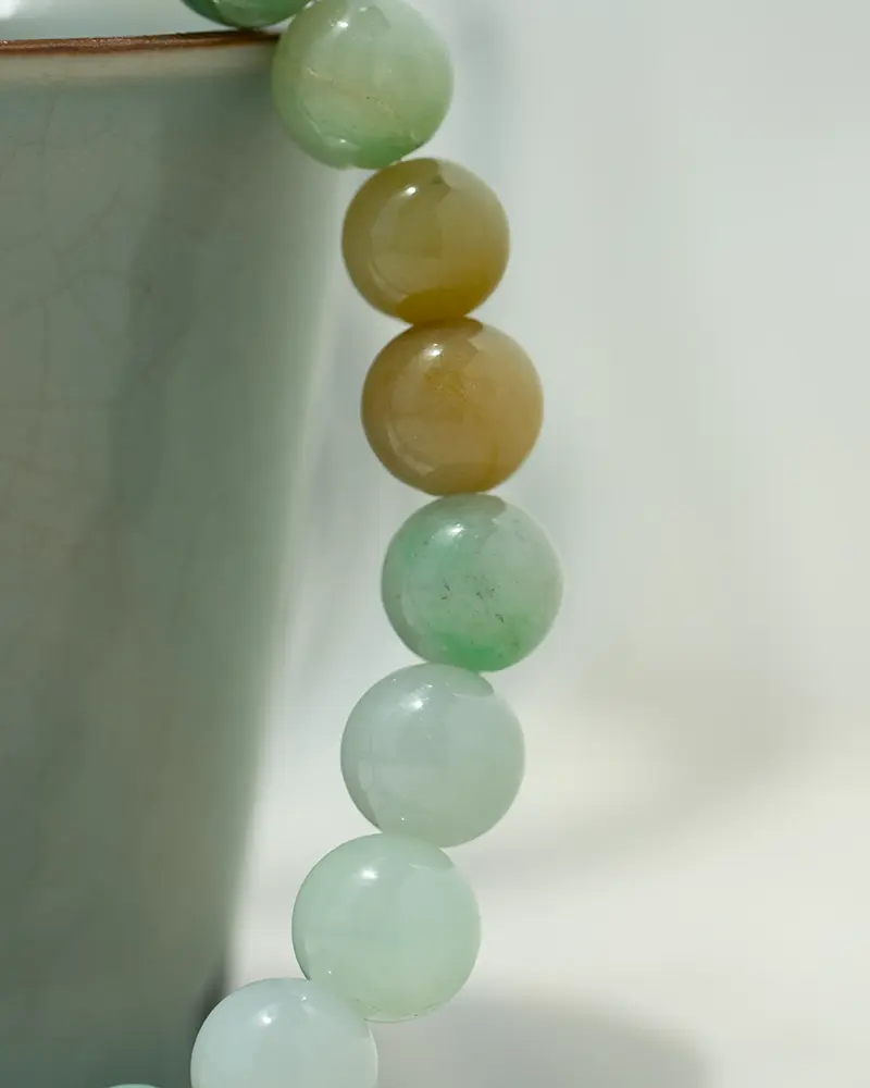 DSC_0565 拷贝 2 A close-up of a beaded jade bracelet draped over the edge of a light green ceramic cup, with beads in soft green and yellowish-brown hues on a neutral background.