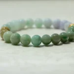 A close-up view of a beaded bracelet made of round, polished green and light-colored stones, resting on a light, textured surface.