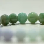 A close-up of a row of polished green jade beads, with a blurred row of lighter-colored beads in the foreground on a light surface.