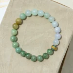 A beaded bracelet made of round, multicolored stones and one decorative gold bead with a floral pattern, displayed on a beige textured surface.