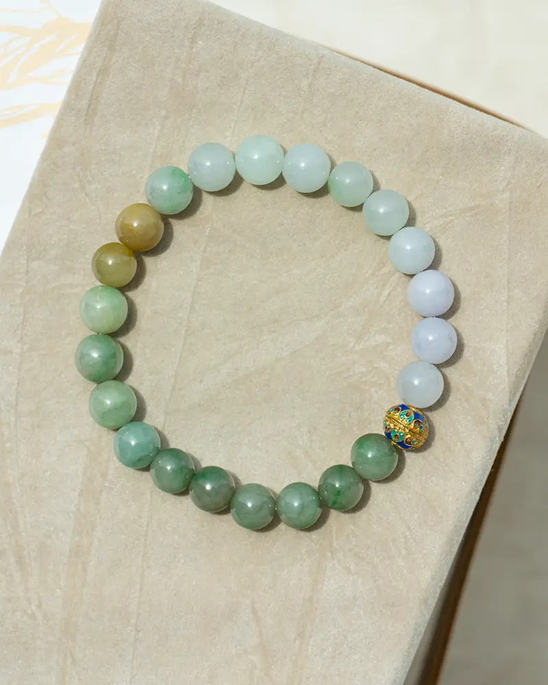 DSC_0569 拷贝 A beaded bracelet made of round, multicolored stones and one decorative gold bead with a floral pattern, displayed on a beige textured surface.