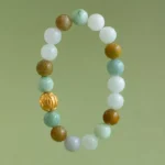 A beaded bracelet with round stones in white, green, and brown hues, and one distinctive gold bead, displayed against a plain light green background.