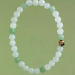 A beaded bracelet made of alternating white and light green round beads, with a single decorative bead featuring gold and red accents, displayed on a light green background.