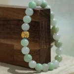 A jade beaded bracelet with one distinctive gold bead is draped over the corner of a wooden block, with a neutral background.