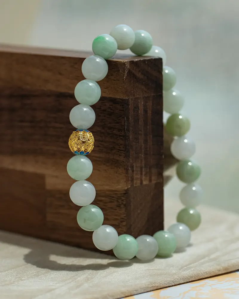 A jade beaded bracelet with one distinctive gold bead is draped over the corner of a wooden block, with a neutral background.