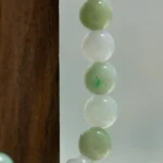 Close-up of a string of round jade beads in light green and white shades, hanging next to a wooden surface with a neutral, blurred background.