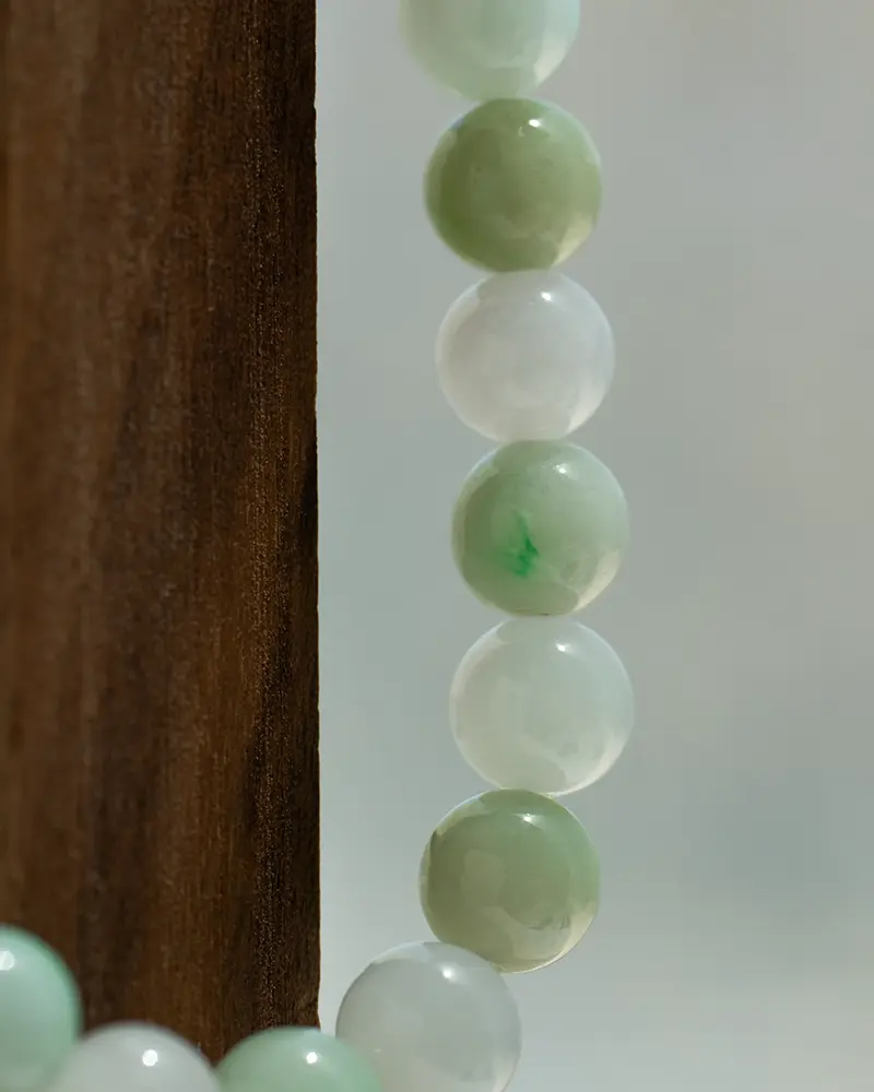 Close-up of a string of round jade beads in light green and white shades, hanging next to a wooden surface with a neutral, blurred background.