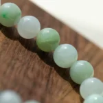 A close-up of a jade beaded bracelet resting on a wooden surface, showing smooth, round beads in varying shades of pale green and white.