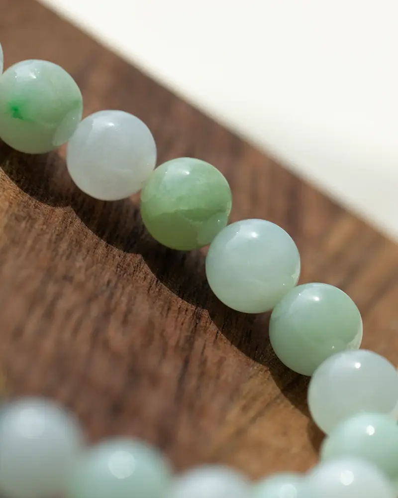 A close-up of a jade beaded bracelet resting on a wooden surface, showing smooth, round beads in varying shades of pale green and white.