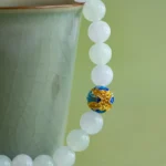 A pale green teacup with a gold rim is next to a jade bead bracelet. One bead is distinct, decorated with blue, gold, and red patterns. The background is also green.