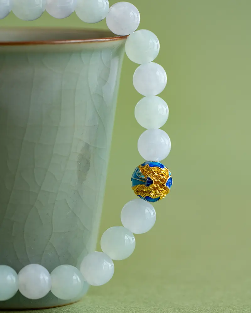 DSC_0584 拷贝 A pale green teacup with a gold rim is next to a jade bead bracelet. One bead is distinct, decorated with blue, gold, and red patterns. The background is also green.