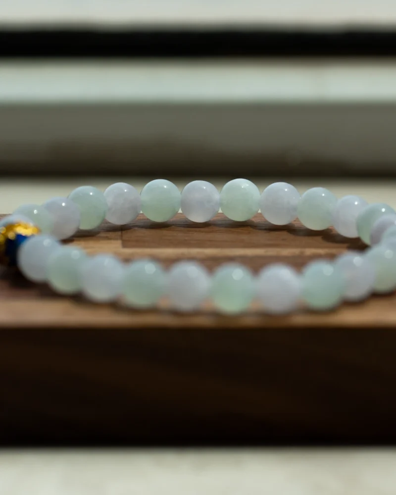 The White and Light Green Jadeite Bead Bracelet — Spherical Beads with Blue Accent Detail rests on a wooden surface, its pale green and white jadeite beads complemented by a single dark blue accent bead near the clasp. The softly blurred background enhances the bracelet’s elegant contrast.