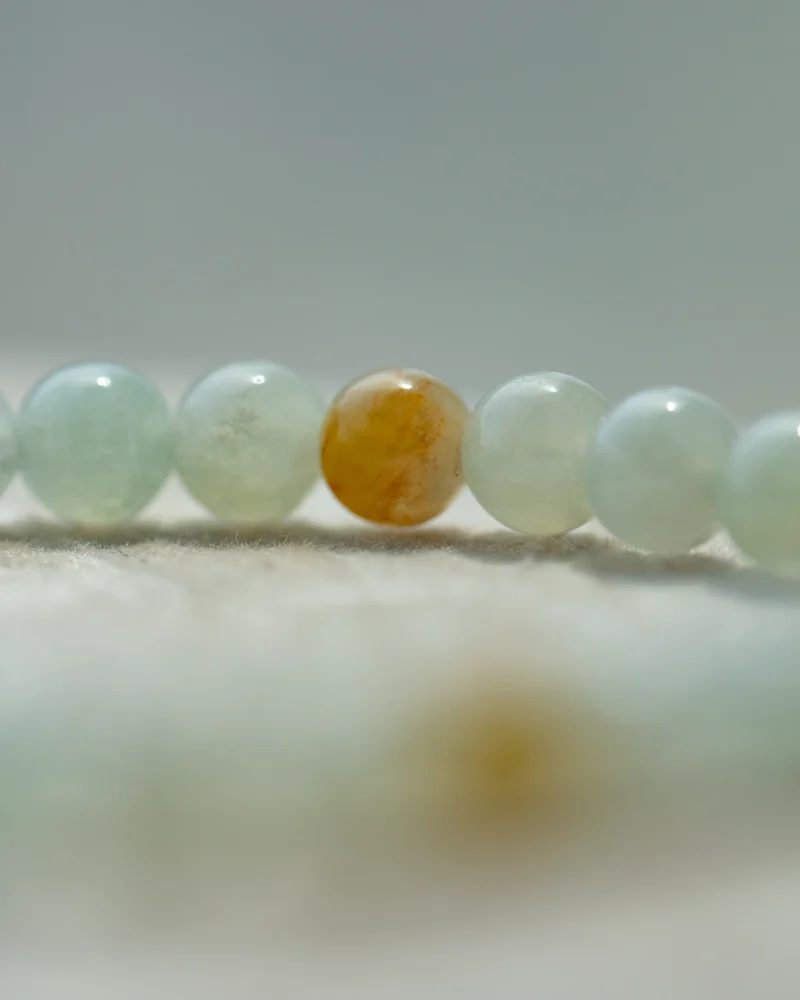 A close-up of the Greenish-White Jadeite Bead Bracelet, featuring predominantly pale green spherical beads accented by a single brownish-yellow bead at the center, displayed on a soft, light surface.
