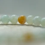 A close-up of a beaded bracelet with mostly pale, translucent beads and one distinct bead that is mottled with yellow and orange tones, resting on a soft, light-colored surface.