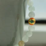 A close-up of a beaded bracelet with pale green and beige round beads, featuring a single ornate bead with red, gold, black, and green patterns, draped over the edge of a light gray ceramic cup.